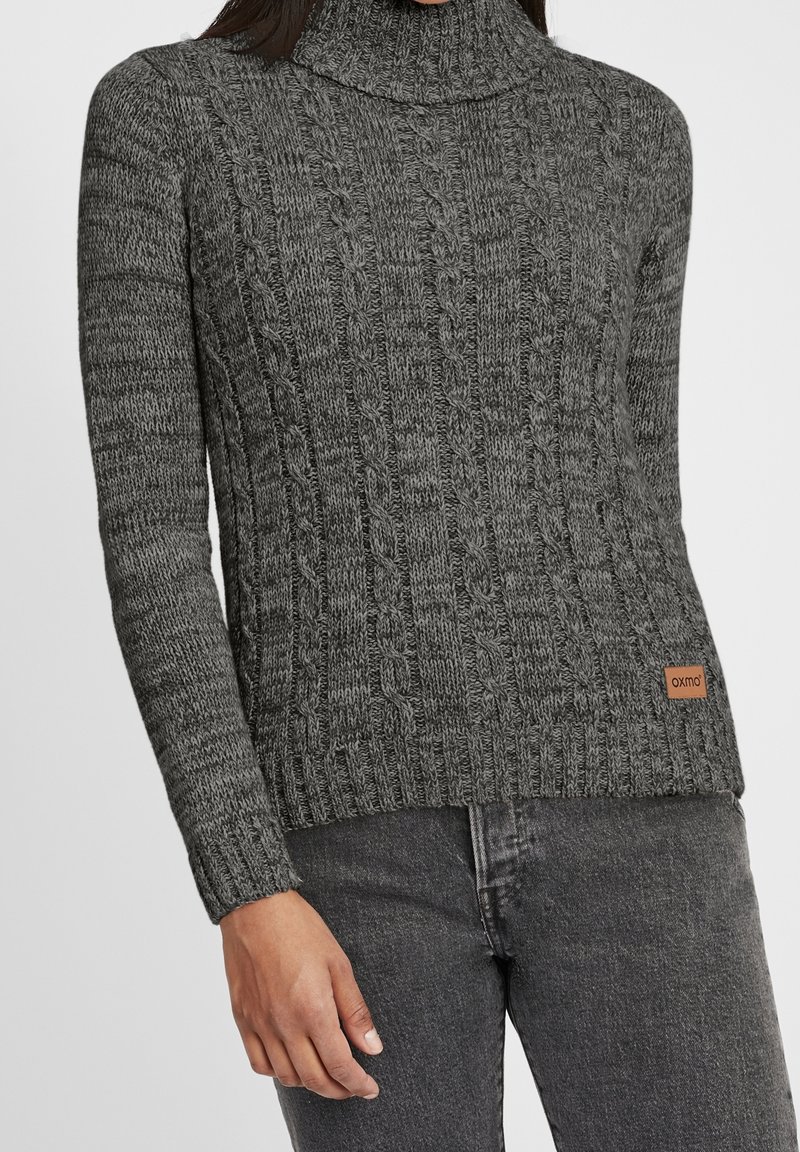 Femme portant un pull à col roulé en maille torsadée gris et un jean gris foncé, debout avec la main gauche détendue le long du corps.