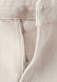 Detailný záber na čiastočne rozopnuté svetlobéžové denimové nohavice s viditeľným kovovým gombíkom a zubami zipsu.