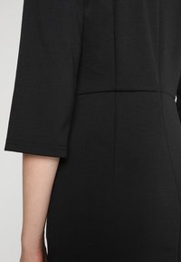 Close-up van de arm en zijkant van een torso van een persoon die een aansluitende zwarte jurk met ellebooglange mouwen en zichtbare naaddetails draagt.