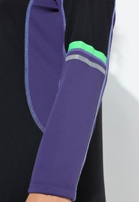 Detailní záběr na fialové a černé tričko s dlouhým rukávem, které má zelený a reflexní proužek poblíž zápěstí na ruce proti bílému pozadí.