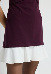 Jupe ajustée bordeaux avec un ourlet froncé blanc, présentant une texture lisse et un détail de poche sur le côté. Vue en gros plan mettant en avant le design.
