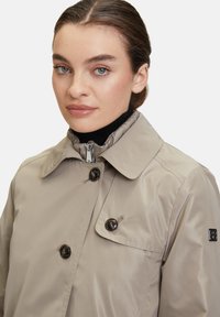 Beige, vattentät trench coat med hög krage, två stora knappar och dragkedja. Slät textur, minimalistisk design och inga mönster.