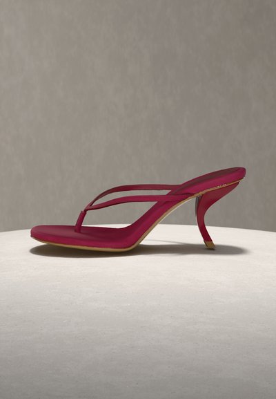 Κόκκινο σανδάλι με τακούνι, σχεδιασμένο με λουράκια και καμπυλωτό τακούνι στιλέτο, τοποθετημένο σε λευκή στρογγυλή επιφάνεια πάνω σε ανοιχτό γκρι φόντο.