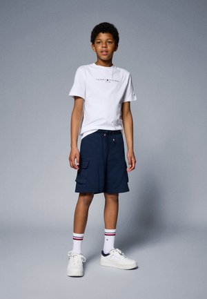 Biały t-shirt z krótkim rękawem z logo, granatowe spodenki cargo z kieszeniami, białe sneakersy z granatowymi akcentami oraz białe skarpetki w czerwone paski.