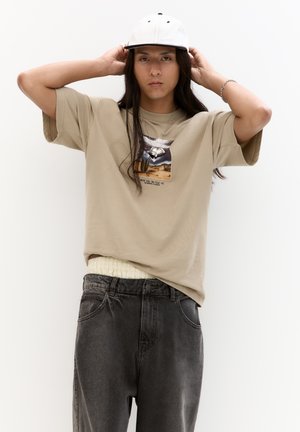 Beige oversized t-shirt med et grafisk print, parret med sorte denim jeans. Modellen bærer en hvid kasket, har langt hår og en sølvhalskæde.