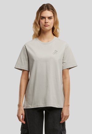 Femme aux cheveux blonds ondulés porte un t-shirt gris clair avec un petit motif cerise sur le côté gauche de la poitrine et un pantalon cargo noir sur fond uni.