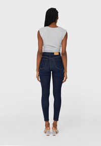 Mujer con el cabello trenzado, vistiendo una blusa sin mangas gris, jeans ajustados de color azul oscuro y sandalias con tacones transparentes, de pie de espaldas sobre un fondo gris.
