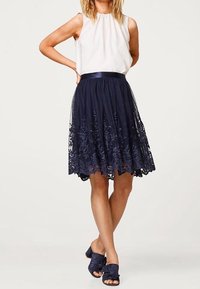 Gonna di pizzo blu navy con ricami floreali, completata da una fascia in satin in vita. Indossata con un top senza maniche bianco e sandali blu con fiocco.