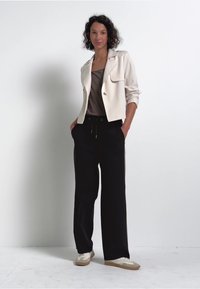 Veste courte crème claire avec col, associée à un pantalon large noir, et des baskets beiges, le tout sur un fond neutre.