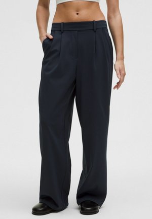 Bukser med vide ben i mørk marineblå, lavet af et glat stof. Har to forreste læg, bæltestropper og sidelommer. Sorte sko fuldender looket.