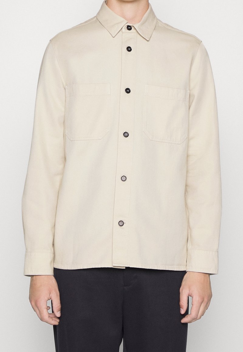 Chemise beige à manches longues avec boutons et deux poches poitrine portée avec un pantalon noir sur une personne debout sur fond neutre.