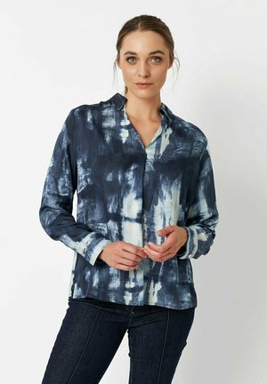 Frau mit zurückgebundenem Haar, die ein langärmliges, knöpfbares Hemd mit marineblauen und weißen Batikmustern sowie dunklen Jeans trägt, vor einem weißen Hintergrund stehend.