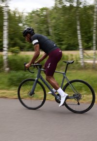 Un cycliste en maillot noir et en shorts bordeaux roule sur un vélo Canyon gris sur un chemin. La verdure environnante et le flou de mouvement suggèrent la vitesse.