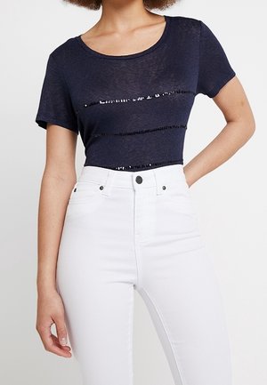 Femme portant un haut ajusté bleu marine à manches courtes avec des bandes horizontales en sequins et un jean blanc taille haute.