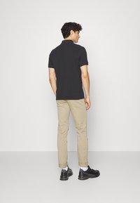 Jaunas vyras stovi atsukęs nugarą, vilkėdamas juodą polo marškinėlį, smėlio spalvos trumpas kelnes, baltas kojines ir juodus batus prieš vienodą foną.