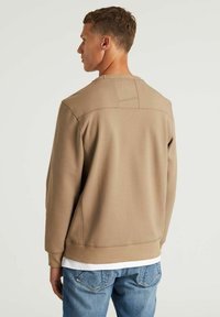 Sudadera beige con cuello redondo y mangas largas. Textura suave, con un panel trasero en contraste y un sutil detalle de costura.