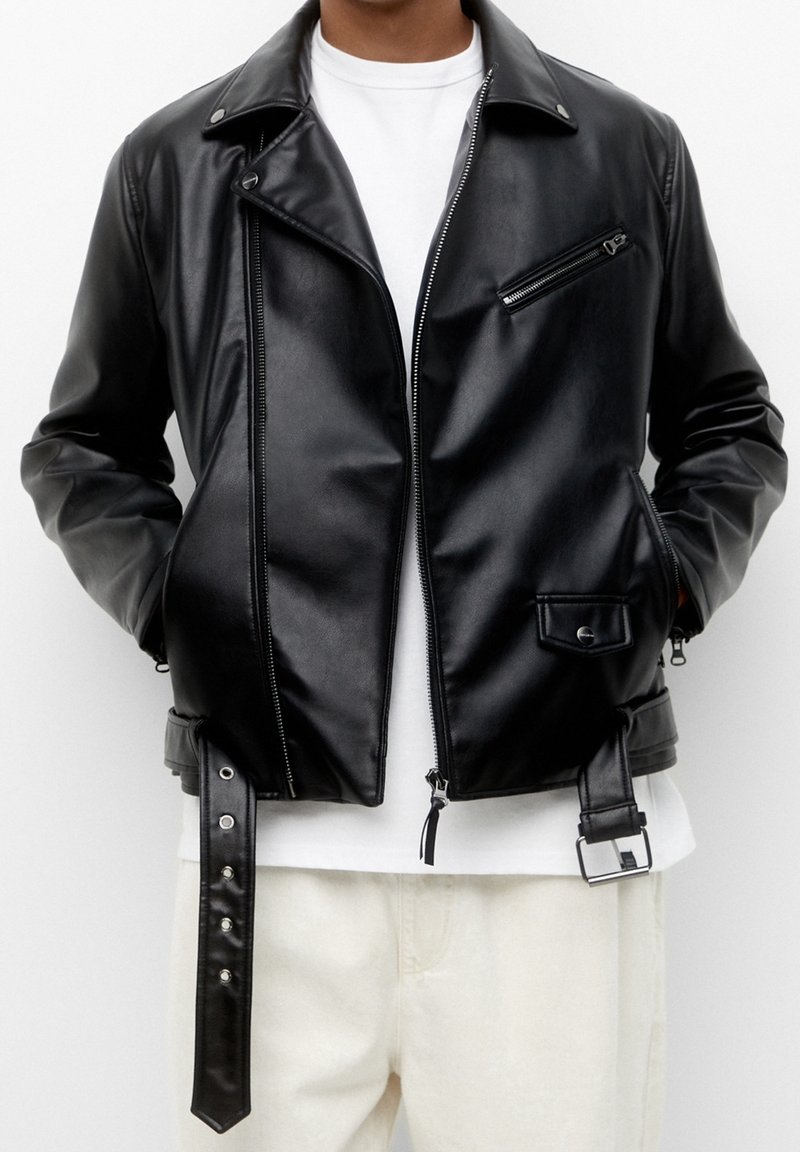 Veste de motard en simili cuir noir avec une fermeture éclair à l'avant, épaulettes, poche poitrine et ceinture ajustable à la taille avec des finitions en métal argenté.