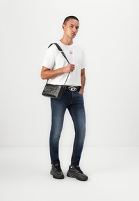 Geantă de umăr neagră, rectangulară, cu logo-ul gravat "DIESEL", purtată cu un tricou alb și blugi de culoare bleumarin. Modelul stă în fața unui fundal alb.