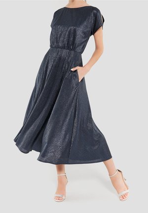 Kobieta ubrana w ciemnoniebieską, połyskującą sukienkę midi z krótkimi rękawami, z ręką w kieszeni, w połączeniu z jasnymi sandałami na wysokim obcasie.