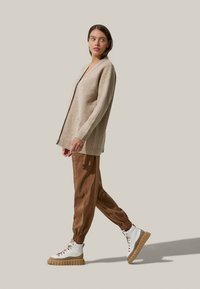 Cardigan beige in lana con scollo a V, polsini a coste e due tasche frontali, abbinato a pantaloni brown a gamba affusolata e sneakers bianche alte con suola spessa.