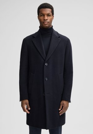 Cappotto di lana blu navy con colletto a punta, tre bottoni frontali e tasche laterali. Texture liscia e design lungo fino al ginocchio. Stile minimalista.