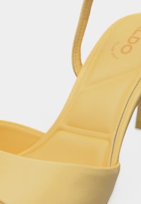 Gros plan d'une sandale à talon haut jaune avec semelle intérieure rembourrée et bride de cheville, montrant une partie de la semelle et du talon.