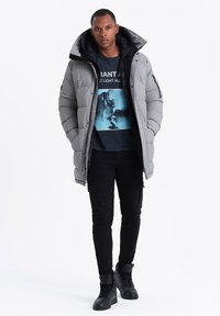 Giacca bombata grigia con colletto alto, caratterizzata da un motivo trapuntato. Indossata sopra una t-shirt nera con grafica e pantaloni cargo, abbinata a sneakers nere.
