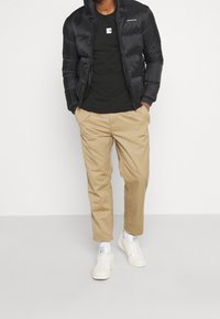 Černá zateplená bunda s kapucí, černé tričko, béžové chinos kalhoty a bílé tenisky. Bunda má zip na přední straně a logo.