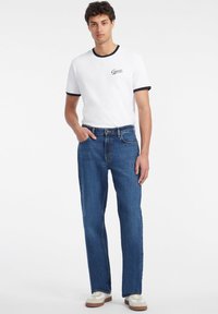 Camiseta blanca de algodón con ribetes negros y logo "Guess", combinada con unos jeans de mezclilla azules, que muestran un ajuste relajado y un diseño clásico.