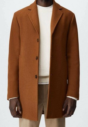 Homme portant un manteau marron croisé simple par-dessus un pull blanc et un pantalon beige, debout devant un fond clair uni.