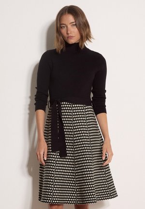 Mujer con cabello hasta los hombros que lleva un top negro de manga larga y una falda línea A con estampado en blanco y negro y cinturón atado.