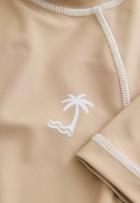 Beige színű, rövid ujjú rash guard, fehér pálmafával ellátott logóval, díszvarrással a galléron és az ujjak szélén.
