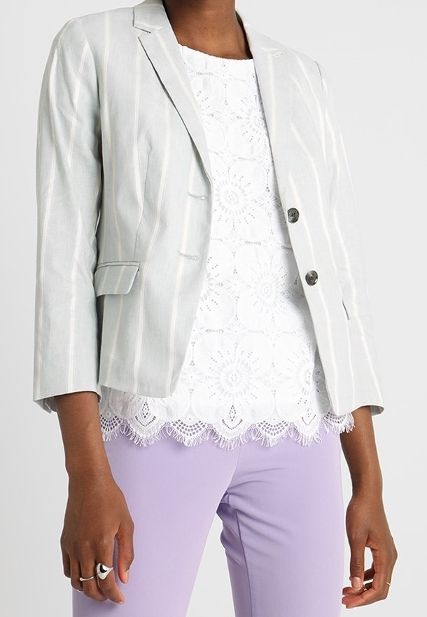 Femme portant un blazer gris clair à fines rayures, un haut en dentelle blanche avec un ourlet festonné, et un pantalon lavande, avec des bagues visibles aux doigts.
