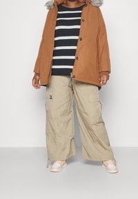 Parka marrón con capucha de pelo sintético, suéter a rayas en azul marino y blanco, pantalones cargo beige y zapatillas blancas con acentos naranjas. Conjunto casual.