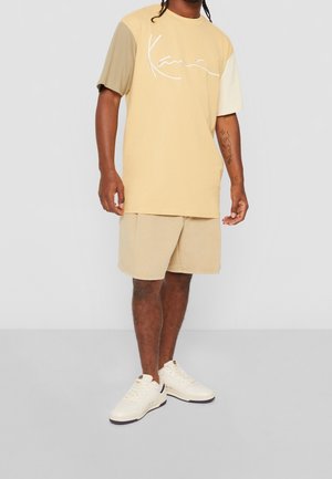 T-shirt oversize couleur beige clair avec manches courtes beiges et logo script blanc ; associé à un short beige et des baskets crème. Texture lisse.