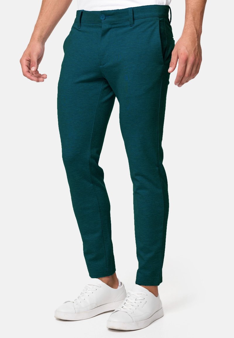 Homme portant un pantalon slim en vert foncé et des baskets blanches, se tenant devant un fond uni.