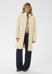 Manteau crème matelassé avec col montant, fermetures à pression et deux poches avant, assorti à un pull beige et un pantalon large bleu foncé.