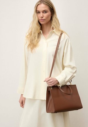 Plavuša u krem košulji i suknji drži veliku smeđu kožnu torbu s remenom na rame na jednoličnoj pozadini.