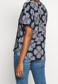 Granatowa bluzka w kwiatowy wzór w odcieniach bieli i niebieskiego. Krótkie rękawy, luźny krój i okrągły dekolt. Lekki materiał, z delikatnym połyskiem.