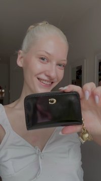 Zwarte lakleren portemonnee met een ritssluiting, gouden hardware en een hartje hanger. Compacte rechthoekige vorm met een gladde textuur.