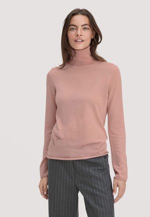 Donna con capelli castani, che indossa un maglione a collo alto rosa polveroso e pantaloni grigi a righe sottili, in piedi davanti a uno sfondo chiaro e neutro.