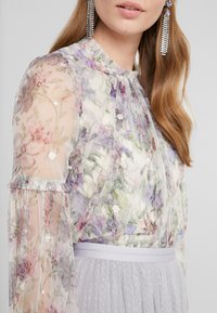 Chemisier transparent à motifs floraux aux tons violets et roses. Il présente des manches à volants et une broderie décorative, associé à une ceinture gris clair.