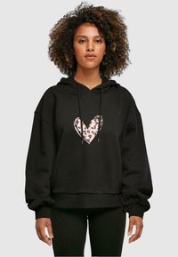 Hoodie noir avec un grand motif de cœur rose à motif léopard. Coupe oversize avec capuche à cordon de serrage et poignets côtelés.