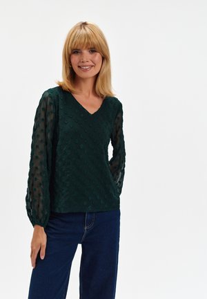 Ciemnozielona góra z teksturowanej tkaniny z wypukłymi wzorami w kropki oraz przezroczystymi długimi rękawami. Dekolt w serek i luźny krój, w zestawieniu z niebieskimi dżinsami.