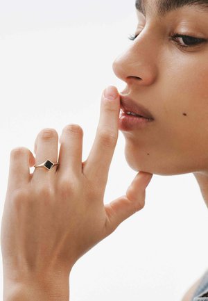 Anillo de oro con una piedra cuadrada negra, llevado en una mano cerca de un rostro de piel suave. Mano posicionada cerca de los labios, dedo tocando la boca.