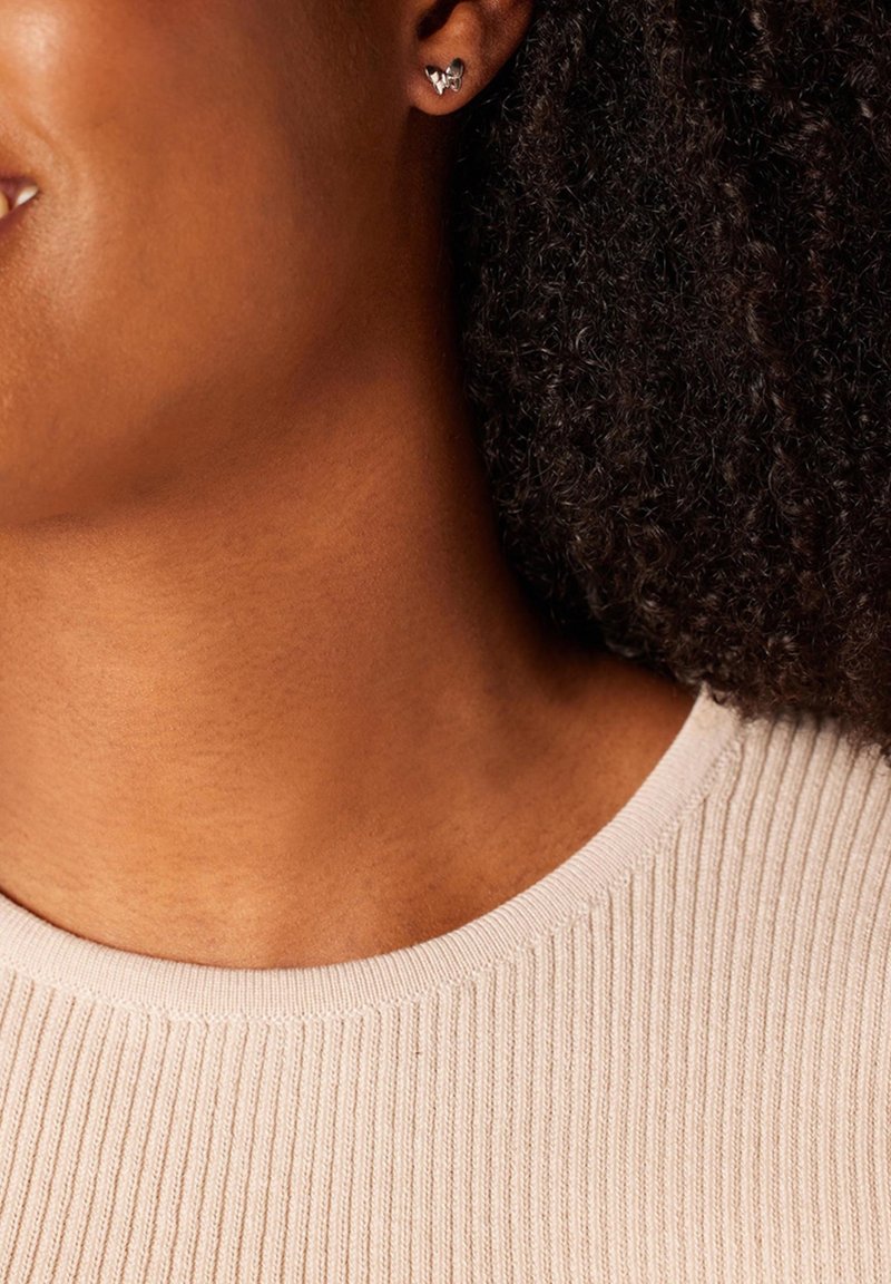 Des boucles d'oreilles en argent en forme de cœur sur une personne aux cheveux foncés et bouclés. Le décolleté est en tissu tricot beige clair et côtelé. Des tons de peau lisses sont visibles.
