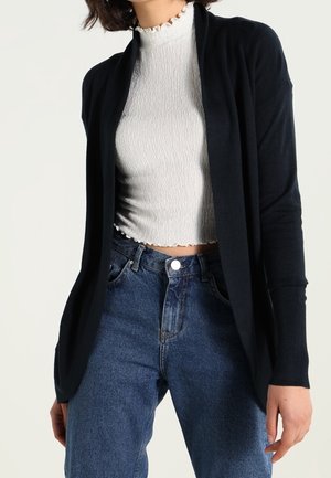 Kvinde iført en kort hvid tekstureret top med flæsekant, mørk åben cardigan og højtaljede blå jeans.