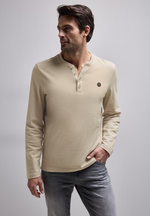 Beige langslijf henley shirt van een textuurstof, met een drieknops placket en een klein logo op de linkerborst. Gepaard met grijze jeans.