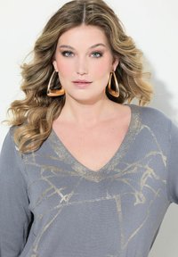 Pull gris à col en V, avec un motif abstrait doré métallique ; modèle avec des cheveux ondulés et de grandes boucles d'oreilles en or. Texture lisse.