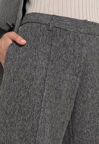 Nära vy av grå byxor i ulltextur med en frontveck, bärs med en beige ribbad topp, handen instoppad i vänster ficka.